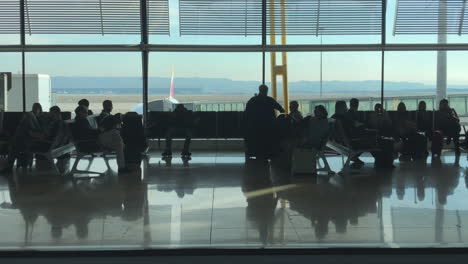 People-on-walkway-rushing-past-the-viewer,-others-sitting-in-airport-terminal,-sliding-dolly-shot,-silhouetted-passengers,-boarding-bridges-and-tarmac-in-background,-travel-and-transportation-concept