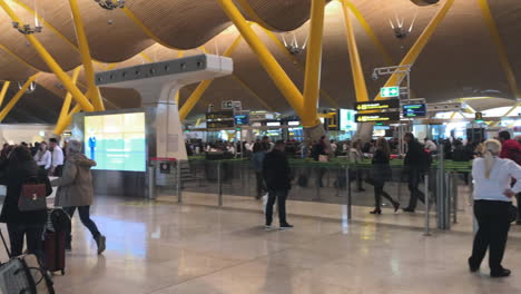 People-rushing-through-terminal-area-of-Madrid-Barajas-International-Airport-and-passing-security-check-towards-their-check-ins,-panning-shot-left-to-right
