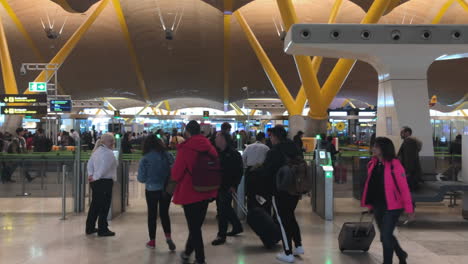 People-rushing-through-terminal-area-of-Madrid-Barajas-International-Airport-and-passing-security-check-towards-their-check-ins,-panning-shot-left-to-right