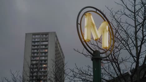 Low-Angle-View-Of-Illuminated-Paris-Metro-Sign-Against-Clouds-and-Building