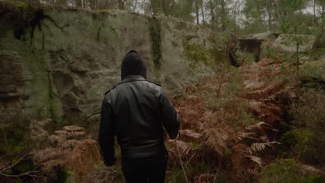 Adult-Hooded-Male-Walking-Through-Forest-Near-Rock-Cliff-Aided-by-Walking-Stick-At-Fontainebleau,-France