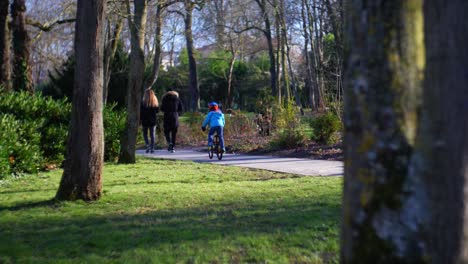 Boy-Riding-His-Bike-On-Path-In-Park-Behind-His-Parents
