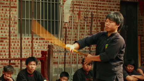 Un-Joven-Hace-Girar-Una-Lanza-De-Kung-Fu-Mientras-Otros-Estudiantes-De-Artes-Marciales-Observan