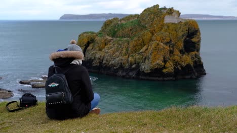 Person-Sitzt-Am-Rand-Einer-Klippe-An-Der-Küste-Nordirlands-Und-Blickt-Auf-Die-Meeresbucht-Mit-Felsbrocken