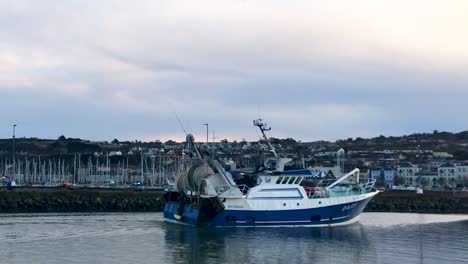 wide-shot-of-fisherboat-passing-by-in-front-of-Howth-Harbour-with-old-town-houses-and-yacht-harbor-in-cozy-sunset-evening-atmosphere