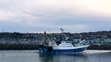 Weitwinkelaufnahme-Eines-Vorbeifahrenden-Fischerbootes-Vor-Dem-Hafen-Von-Howth-Mit-Alten-Stadthäusern-Und-Yachthafen-In-Gemütlicher-Abendstimmung-Bei-Sonnenuntergang