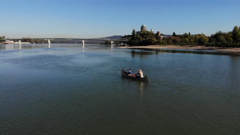 Aerial-view-of-two-males-canoeing-upstream-on-the-Danube-river-at-Esztergom,-Hungary-in-the-background-with-the-Basilica