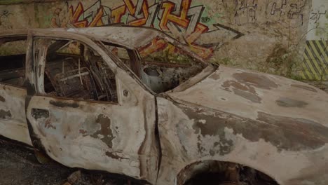 POV-View-Of-Burnt-Out-Wreckage-Of-Car-In-Abandoned-Warehouse-With-Graffiti-On-Walls