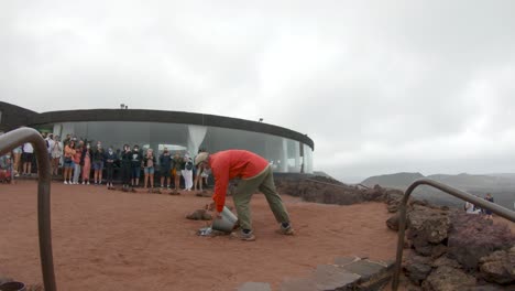 Vulkanischer-Geysir-Im-Nationalpark-Timanfaya,-Lanzarote-–-Touristen-Beobachten,-Wie-Ein-Mann-Einen-Eimer-Wasser-In-Das-Loch-Auf-Dem-Boden-Schüttet-Und-Dann-Als-Heißer-Dampf-An-Die-Oberfläche-Zurückspritzt