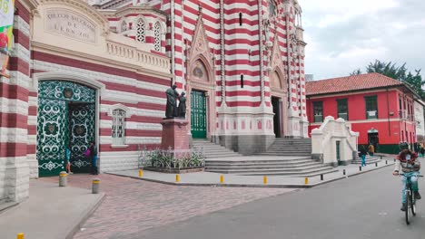 A-man-on-a-bike,-tourists-walking-and-a-motorcycle-passing-on-a-street-next-to-a-Cathedral-in-the-old-town-of-Bogotá,-Colombia