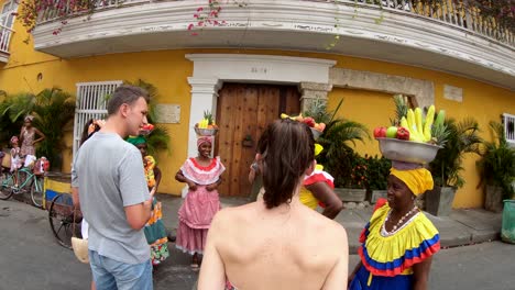 Eine-Frau-Nähert-Sich-Einer-Gruppe-Von-Palenqueras,-Die-Bunte-Kleider-Tragen-Und-Obstschalen-über-Ihren-Köpfen-Balancieren,-Während-Sie-Auf-Einer-Straße-Der-Altstadt-Von-Cartagena-De-Indias-Stehen