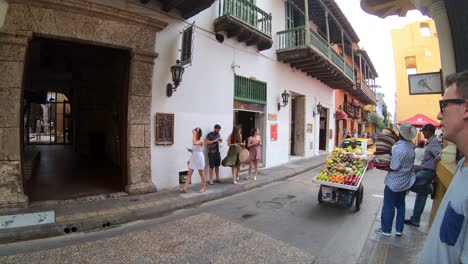 Mehrere-Touristen-Gehen-Eine-Straße-In-Cartagena-(Indien)-Entlang,-Während-Ein-Mann-Sein-Wagenrad-Voller-Verkaufsfertiger-Früchte-Schiebt