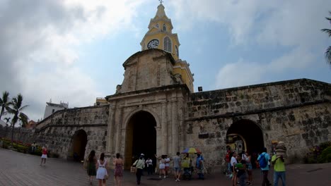 A-group-of-women-and-tourists-are-walking-close-to-an-old-arc,-wall-and-clock-tower-that-is-the-entrance-of-the-old-town-of-Cartagena-de-Indias,-Colombia