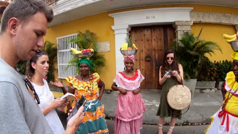 Tourists-are-standing-and-talking-with-palenqueras-who-are-wearing-colorful-dresses-and-balancing-a-fruit-bowl-over-their-heads-in-the-old-town-of-Cartagena-de-Indias,-Colombia