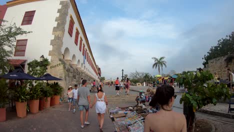 Eine-Frau-Mit-Einem-Kleid-Geht-Hinter-Ein-Paar-Touristen-Her,-Die-Sich-In-Der-Altstadt-Von-Cartagena-De-Indias,-Kolumbien,-An-Den-Händen-Halten