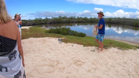 Several-tourists-are-taking-a-picture-of-one-tourist-holding-a-bag-and-standing-on-white-sand-near-a-small-lake-of-an-island-of-the-Galapagos-islands