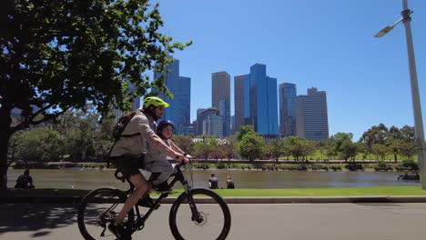 Papá-E-Hijo-Andan-En-Bicicleta-A-Lo-Largo-Del-Río-Yarra-Y-Como-Telón-De-Fondo-La-Ciudad-De-Melbourne