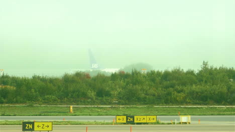 Aircraft-taxiing-behind-tree-line,-Airport-taxiway-signs-on-foreground