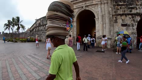 Un-Hombre-Con-Camisa-Verde-Lleva-En-La-Cabeza-Decenas-De-Sombreros-Para-Vender-Cerca-De-Un-Antiguo-Arco-Y-Muro-De-Piedra-Que-Es-La-Entrada-De-Una-Plaza-En-El-Casco-Antiguo-De-Cartagena-De-Indias
