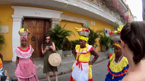 Two-tourists-are-standing-and-speaking-with-three-palenqueras-who-are-wearing-colorful-dresses-and-balancing-fruit-bowls-over-their-heads-on-the-old-town-of-Cartagena-de-Indias,-Colombia