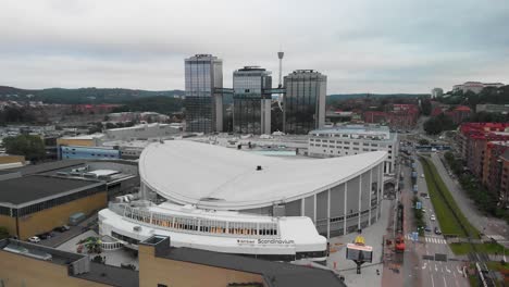 Aerial-view-of-the-famous-concert-arena-Scandinavium-in-Gothenburg,-Sweden,-with-the-luxurious-hotel-Gothia-towers-in-the-background-and-also-a-part-of-the-amusement-park-Liseberg-showing
