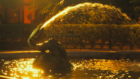 Estatua-En-Una-Fuente-De-Agua-Rociando-Un-Chorro-De-Agua-En-La-Piscina-Por-La-Noche-Con-Una-Luz-Dorada-Brillando-En-La-Superficie