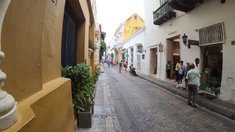 In-a-street-of-Cartagena-de-Indias,-a-man-and-seller-is-carrying-a-cart-wheel-of-fruits-while-several-tourists-are-walking-close