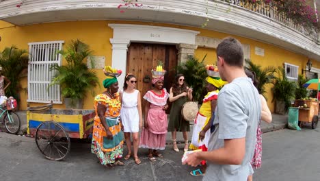 Eine-Gruppe-Von-Touristen-Spricht-Mit-Mehreren-Palenqueras,-Die-Bunte-Kleider-Tragen-Und-Obstschalen-über-Ihren-Köpfen-Balancieren,-Auf-Einer-Straße-Der-Altstadt-Von-Cartagena-De-Indias,-Kolumbien