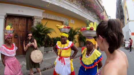 A-group-of-palenqueras-with-colorful-dresses-are-balancing-fruit-bowls-on-their-heads-while-talking-with-two-women-who-are-doing-tourism-in-the-old-town-of-Cartagena-de-Indias,-Colombia