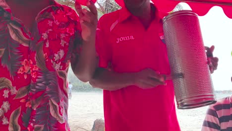 Un-Hombre-Vestido-Con-Una-Camisa-Hawaiana-Está-Junto-A-Un-Hombre-Que-Toca-Canciones-De-Guacharaca-Y-Vallenato-En-Una-Playa-De-Cartagena-De-Indias,-Colombia