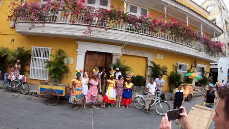 A-man-is-using-his-phone-to-take-a-picture-of-palenqueras-and-more-tourists-next-to-a-yellow-house-and-balconies-with-flowers-in-the-old-town-of-Cartagena-de-Indias