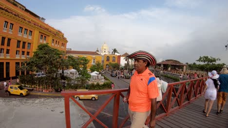 Un-Hombre-Con-Una-Camisa-Naranja-Y-Un-Sombrero-Lleva-Un-Helado-Mientras-Camina-Por-Un-Puente-Rojo-Del-Casco-Antiguo-De-Cartagena-De-Indias,-Colombia