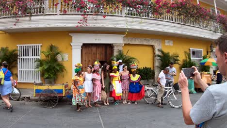A-man-is-taking-a-picture-of-three-palenqueras-and-three-women-next-to-a-yellow-building-with-balconies-and-flowers-in-the-old-town-of-Cartagena-de-Indias,-Colombia