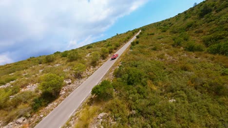 Vista-Aérea-Cinematográfica-De-Un-Mustang-Rojo-Clásico-Conduciendo-Por-Una-Carretera-Panorámica-A-Través-De-Las-Colinas-De-España.