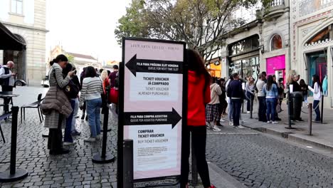 Gente-Haciendo-Cola-En-Una-Calle-Adoquinada-Con-Carteles-En-Un-Día-Soleado