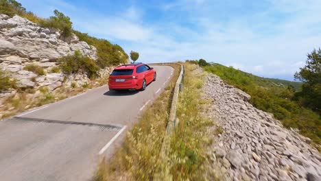 Antena-FPV-Siguiendo-Un-Coche-Rojo-Conduciendo-Por-Una-Carretera-Panorámica-En-El-Campo-Español