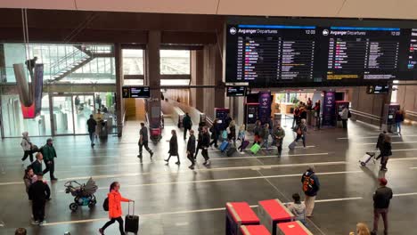 Schwenkaufnahme-Des-Inneren-Des-Osloer-Hauptbahnhofs-Mit-Vielen-Reisenden,-Die-Mit-Gepäck-Unterwegs-Sind,-Und-Leuten,-Die-Auf-Die-Großen-Bildschirme-Schauen,-Um-Reiseinformationen-Zu-Erhalten