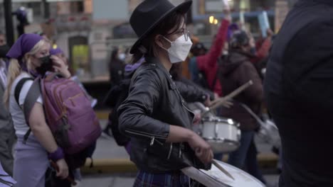 A-woman-wearing-a-white-mask-and-a-black-hat-is-playing-drums-during-the-International-Women's-day-in-Quito,-Ecuador
