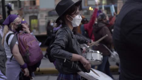 Una-Mujer-Con-Una-Máscara-Blanca-Y-Un-Sombrero-Negro-Toca-La-Batería-Durante-El-Día-Internacional-De-La-Mujer-En-Quito,-Ecuador.