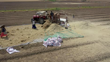 Aerial-drone-rotating-zoom-out-shot-over-farm-workers-pushing-bundles-of-wheat-in-a-thresher-machine-to-separate-grain-from-husk-straw-in-Rajkot,-Gujarat,-India