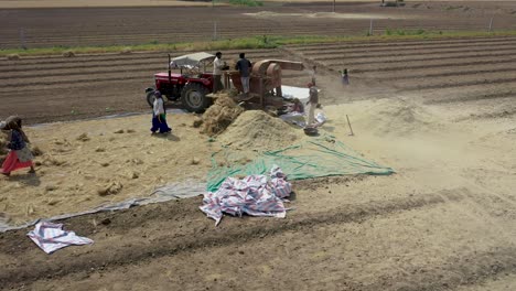 Avión-Teledirigido-Con-Zoom-De-Alejamiento-Giratorio-Sobre-Trabajadores-Agrícolas-Empujando-Manojos-De-Trigo-En-Una-Máquina-Trilladora-Para-Separar-El-Grano-De-La-Paja-De-Cáscara-En-Rajkot,-Gujarat,-India