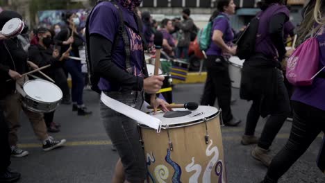 Un-Grupo-De-Mujeres-Vestidas-De-Púrpura-Tocan-Tambores-Mientras-Marchan-Durante-La-Protesta-En-El-Día-Internacional-De-La-Mujer-En-Quito,-Ecuador.
