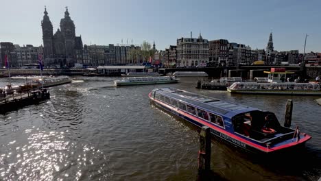 A-congested-dock-outside-the-Amsterdam-Central-station,-as-cruise-boats-navigate-their-arrival-and-departure-on-a-busy-beautiful-sunny-morning,-Netherlands