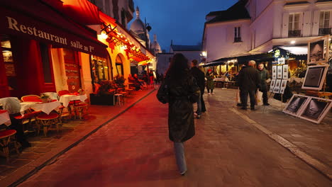 Young-woman-walking-down-a-busy-street-with-restaurants-and-art-shops,-in-Paris-near-Montmartre,-at-night