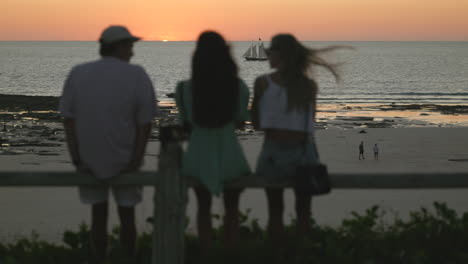 Freunde-Unterhalten-Sich,-Während-Sie-Einem-Segelboot-Zusehen,-Wie-Es-Am-Strand-In-Richtung-Sonnenuntergang-Fährt