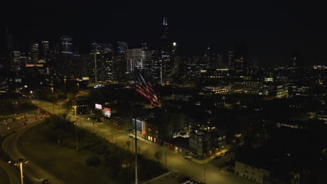 Aerial-view-still-in-front-of-a-waving-US-Flag-in-Chicago-city,-nighttime-in-Illinois,-USA