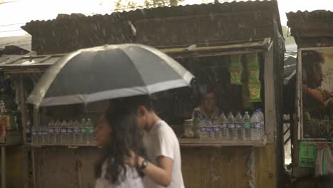 People-and-young-couple-walking-in-rain-with-umbrella,-poor-old-shopkeeper-looking-at-them,-monsoon-season,-slow-motion-shot-of-rain-drops
