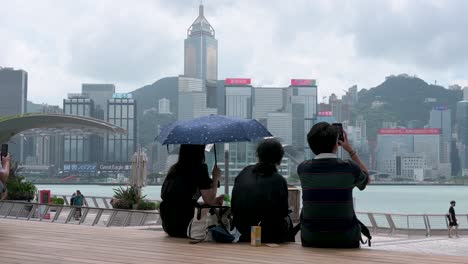 Junge-Touristen-In-Schwarzer-Kleidung-Machen-Fotos-Mit-Ihren-Smartphones-Und-Genießen-Dabei-Den-Blick-Auf-Den-Hafen-Und-Die-Skyline-Von-Hongkong