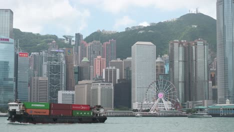 Blick-Auf-Die-Uferpromenade-Des-Victoria-Harbour,-Während-Ein-Frachtschiff-Mit-Containern-Vor-Der-Skyline-Von-Hongkong-über-Den-Rahmen-Fährt