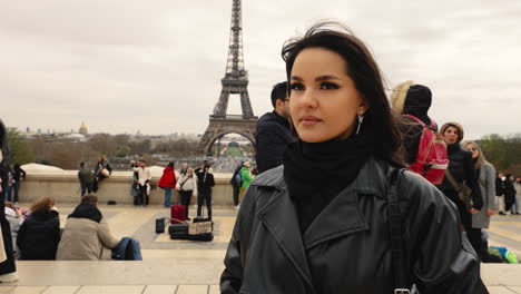 Hermosa-Morena-Parada-Frente-A-La-Torre-Eiffel,-Mirando-A-La-Cámara,-Primer-Plano-Cinematográfico-En-Cámara-Lenta,-Gente-En-El-Fondo